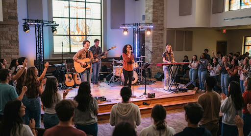 A wide shot of a youth ministry worship band playing on a small stage. Modern lighting design with subtle stained glass motifs in the background. Energetic yet reverent mood. Focus on the connection between the musicians and the congregation.