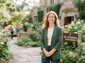 A welcoming female youth pastor in her 30s, smiling warmly, wearing a casual but professional outfit. Photographed outdoors near the church gardens with soft, bright natural daylight. The mood is approachable, joyful, and community-focused.