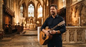 A thoughtful male worship pastor in his 40s, holding a guitar subtly in the frame. Wearing a tailored button-down shirt. Photographed inside the cathedral near the altar with warm, dramatic lighting highlighting the architectural details in the background.