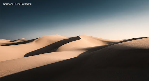 An abstract, stylized shot of rolling desert dunes under a dramatic, high-contrast sky. The sand has a soft, golden hue, while deep shadows create strong geometric shapes. The image feels vast, minimalist, and slightly ethereal, evoking themes of journey and wilderness. Contemporary editorial style.